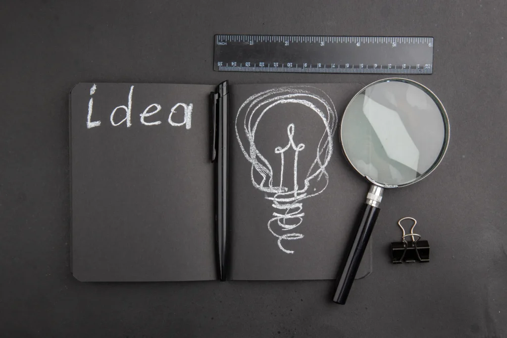 Taccuino nero aperto con la scritta Idea, disegno di una lampadina a gesso, penna, righello e lente d'ingrandimento.