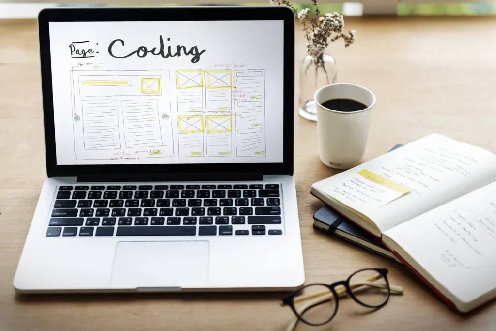 Laptop su scrivania con wireframe di una pagina web a schermo, taccuino, occhiali e tazza di caffè.