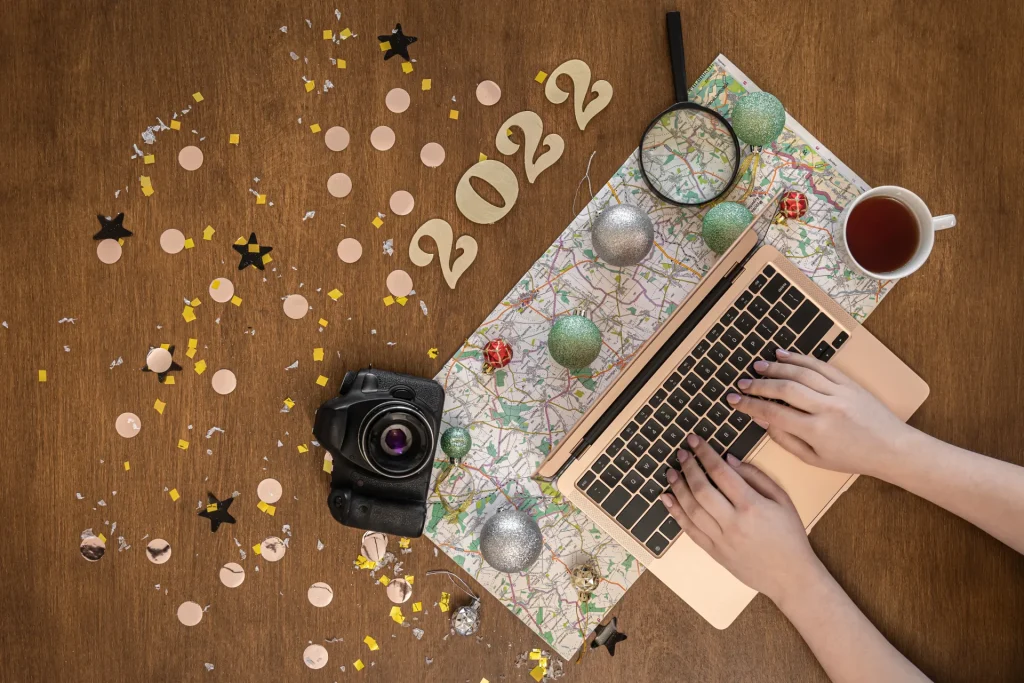 Mani su laptop con fotocamera, mappa, tazza di tè e decorazioni festive con numero 2022 su scrivania in legno.